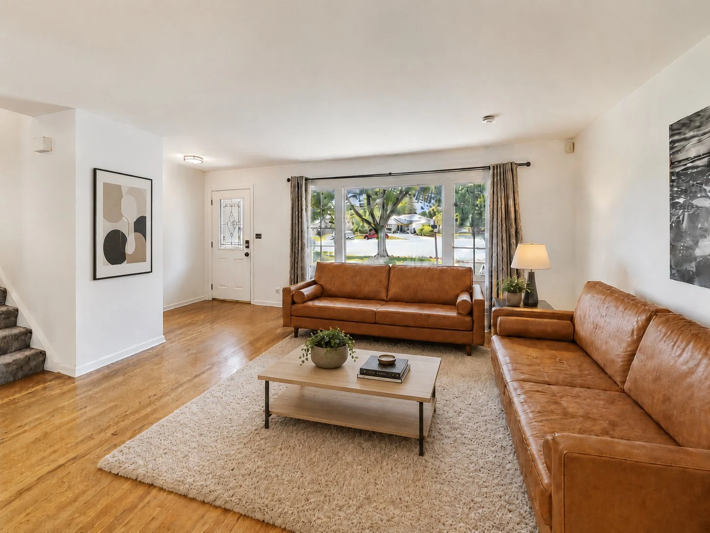 Sun-filled main-level living room with two tan leather sofas, a light wood coffee table, a soft area rug over hardwood floors, framed wall art, and a large picture window looking out to the tree-lined front yard. Stairs lead to the upper level and the front entry is just beyond.