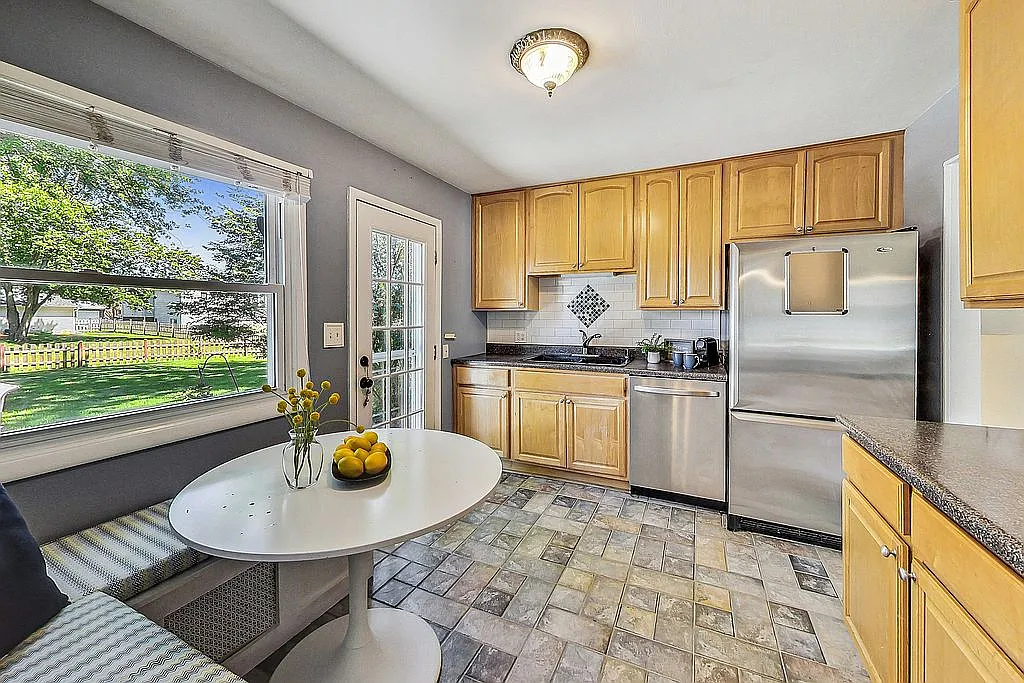 Bright kitchen with light wood cabinetry, stainless-steel refrigerator and dishwasher, tile flooring, a tile backsplash, and a sunny window over the sink overlooking the fenced backyard.