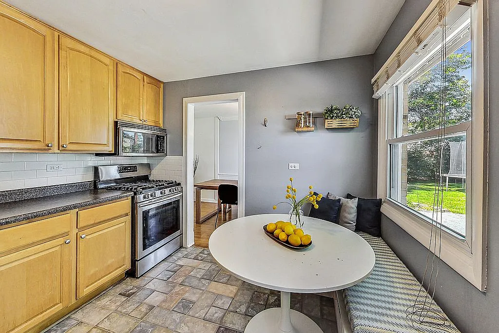 Kitchen view showing a stainless-steel gas range and microwave, light wood cabinets, a window bench with throw pillows beside a sunny window, and a doorway through to an adjoining room.