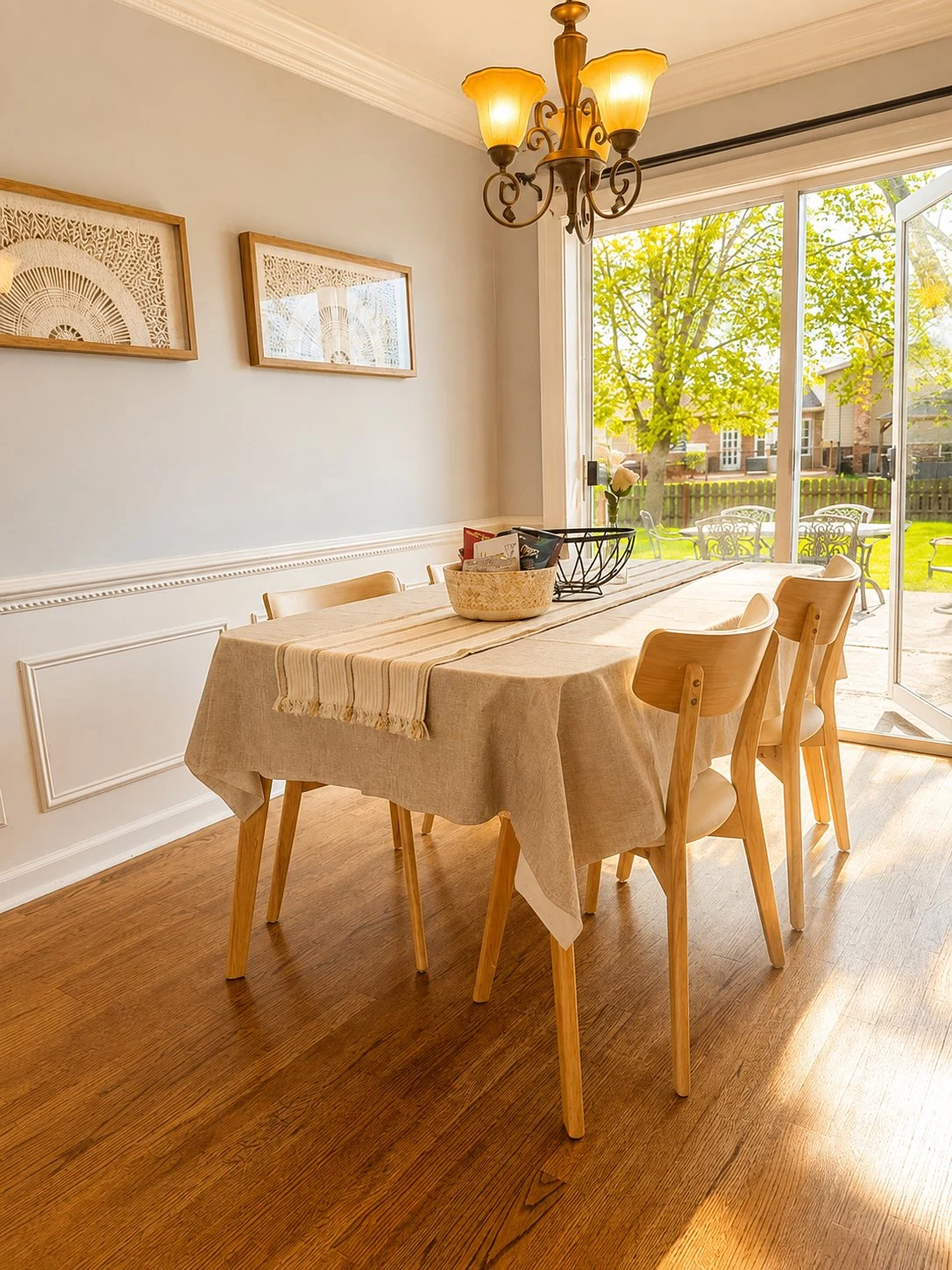 Dining room with a wood table and six chairs dressed with a linen runner, hardwood floors, a brass chandelier, framed wall art, and a wall of sliding glass doors opening to the backyard patio.
