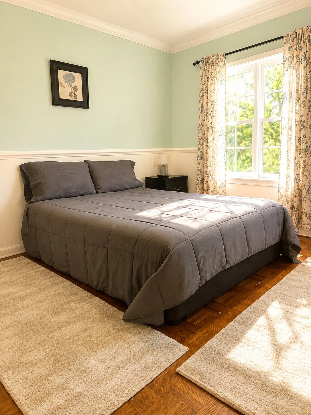 Second bedroom with soft sage-green walls, a queen bed with charcoal bedding, hardwood floors, layered woven area rugs, floral curtains, and a framed botanical print.