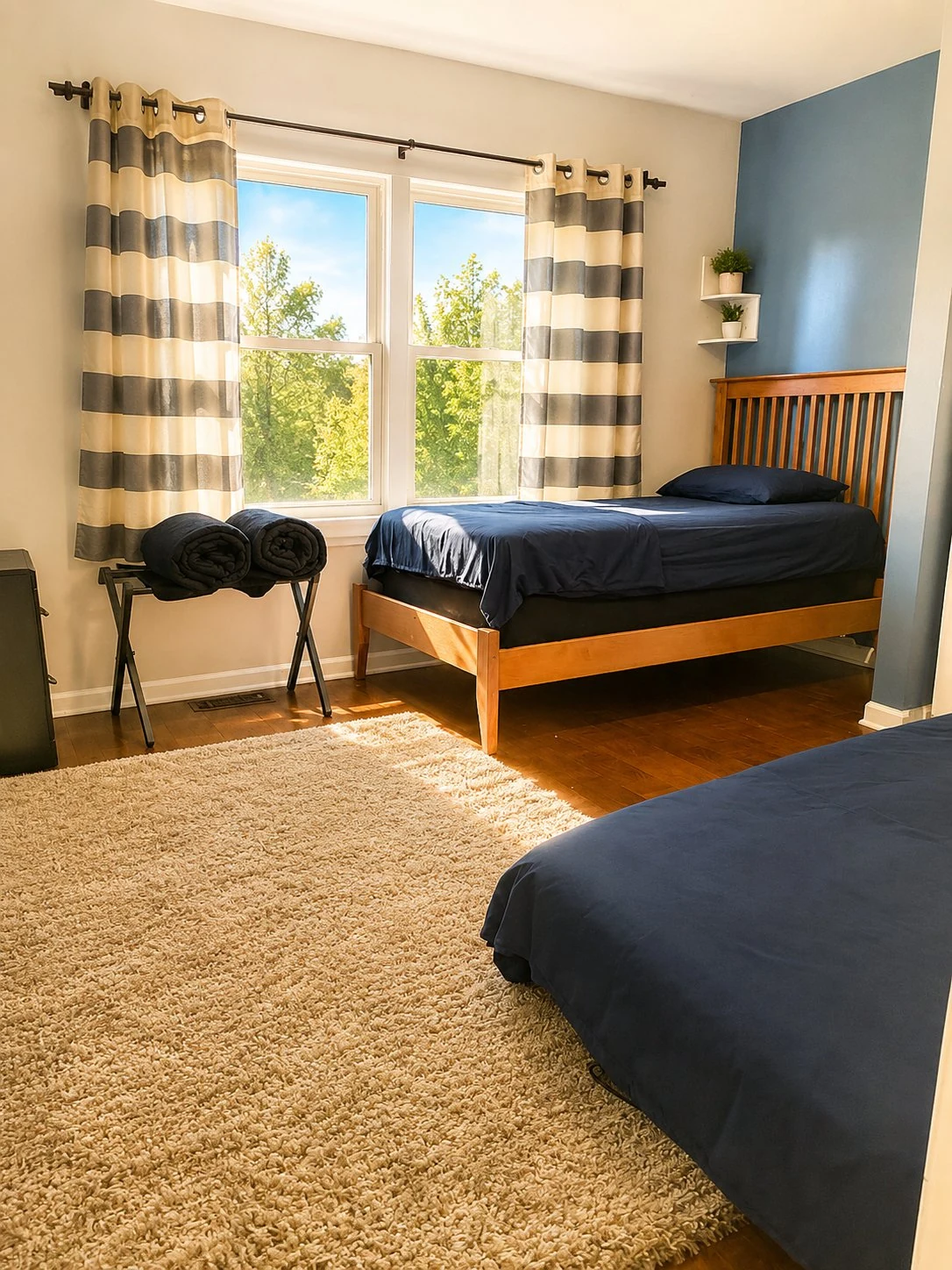 Third bedroom with two beds dressed in navy bedding, gray-and-white striped curtains, hardwood floors, a woven area rug, and a small wall shelf with greenery.