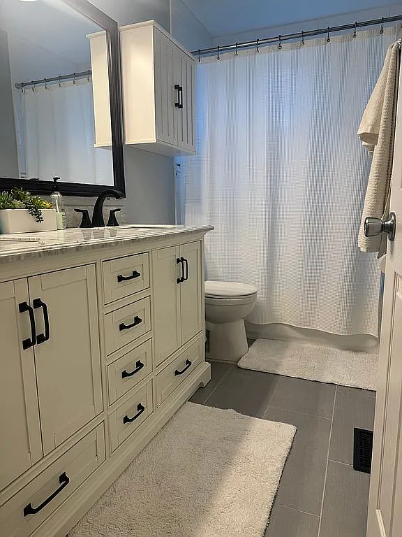 Full bathroom with a white shaker-style vanity, stone countertop, matte black hardware, a framed mirror, gray plank tile flooring, and a shower/tub behind a white waffle-weave curtain.