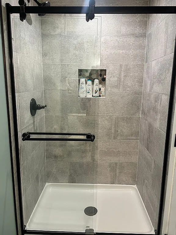 Walk-in shower with sliding glass doors, large gray tile surround, a built-in shampoo niche, matte black fixtures, and a clean white shower base.