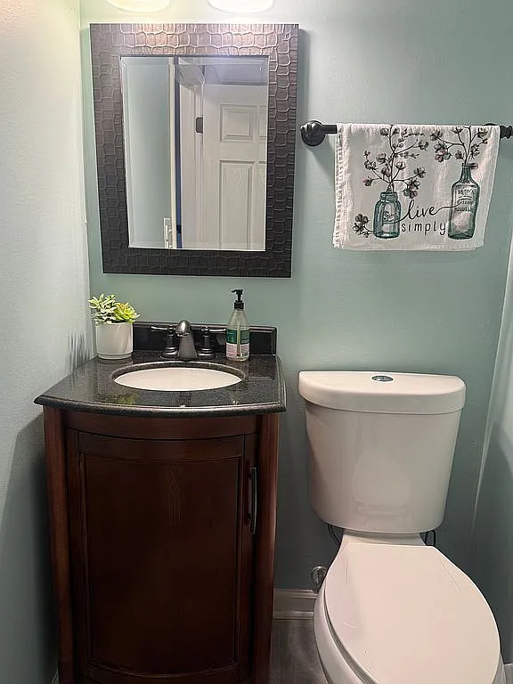 Powder room with a dark wood single-sink vanity, granite countertop, a framed mirror with textured tile detail, soft blue-green walls, and a decorative hand towel.