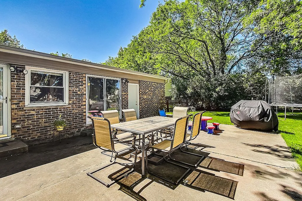Backyard concrete patio with a six-seat outdoor dining set, a covered grill, kids' stools, brick exterior wall with sliding glass doors, and a trampoline visible on the green lawn beyond.