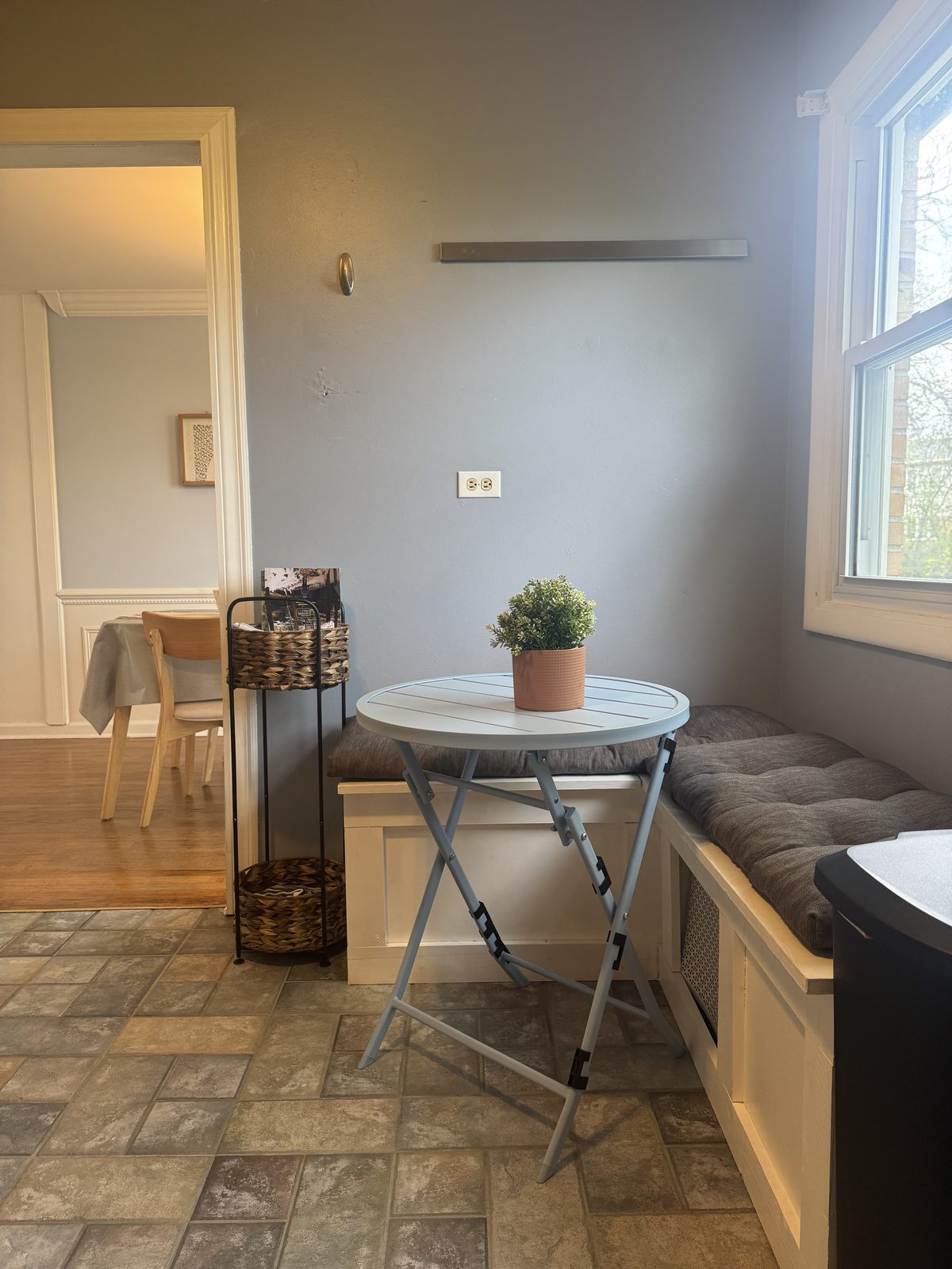 Sunny breakfast nook with a built-in white window seat, gray tufted cushion, light blue bistro table, and a pass-through view to the dining room.