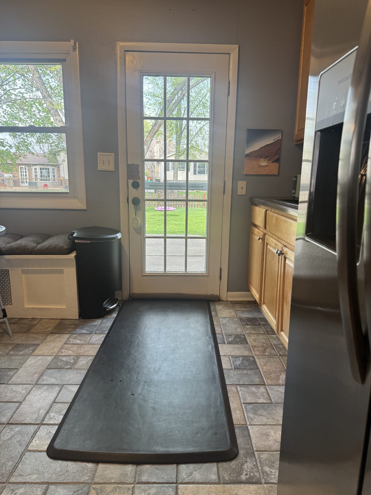 Bright eat-in kitchen with stainless steel side-by-side refrigerator, light wood cabinetry, a built-in window bench, and a glass-paneled door that opens to the patio for grilling.