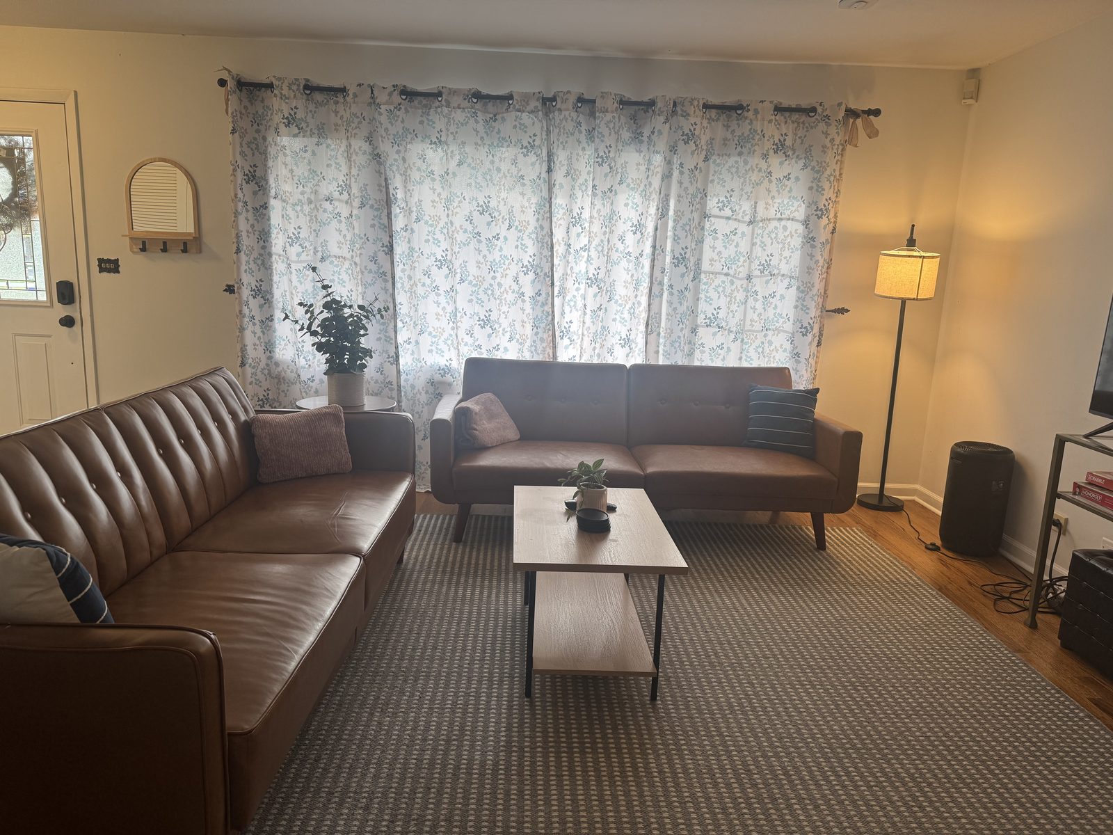 Sun-filled main-level living room at 318 W Hackberry with two brown leather sofas, a wood coffee table, an area rug, and floral curtains over a wide picture window letting in natural light.