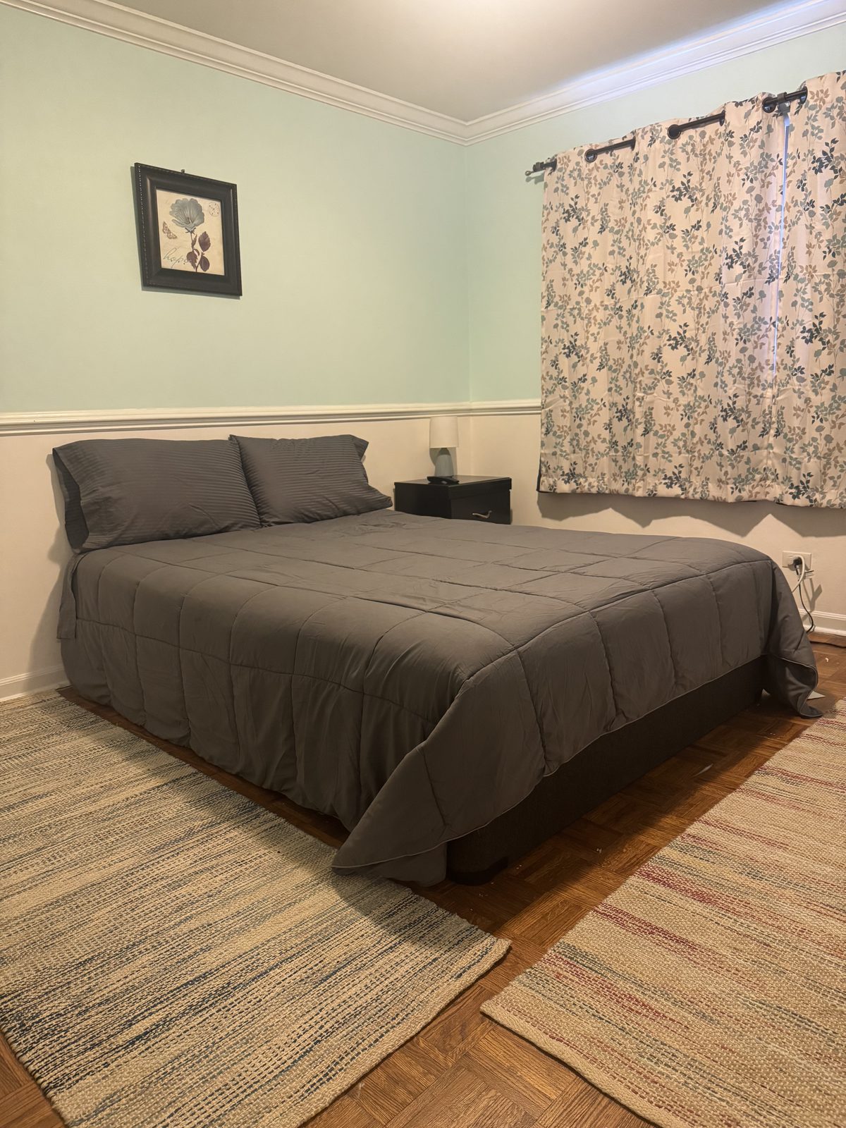 Second upstairs bedroom with soft sage-green walls, a queen bed with charcoal bedding, original hardwood parquet floors, layered woven area rugs, and floral curtains.
